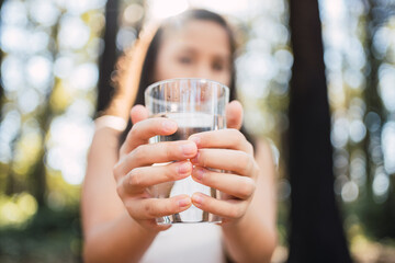 Close-up of a girl's hands holding a glass of pure mineral water, offering to the camera - A pretty little girl who recommends a healthy lifestyle habit, drinking clean water to refresh herself.