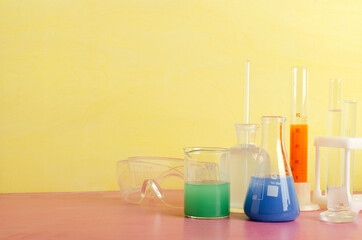 Closeup of laboratory glassware, flakes, protective plastic glasses on the bright pink table against yellow wall.Empty space