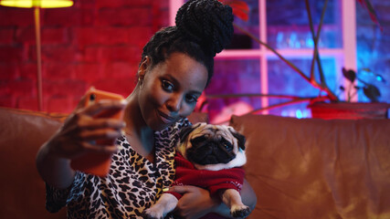 Beautiful african woman taking selfie with dog on sofa at home