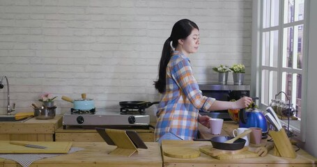 slow motion asian japanese female filling mug with hot water while brewing beverage in kitchen. beautiful lady standing in home cooking place against city view from window. thristy woman drinking.