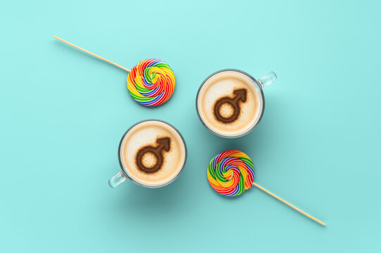 Two cups of coffee with symbols of mars on whipped milk foam and couple round LGBT rainbow lollipops on pastel cyan background. Concept gay love. Flat lay, top view