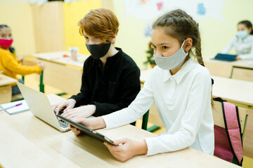 Obraz premium Students in medical masks sit at the table and take part in an online lesson using gadgets