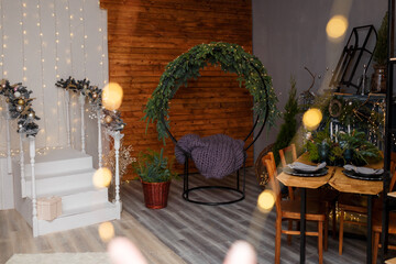 Rustic style Christmas festive decorations, fireplace with firewood, table, chairs, circle sitting swing with fresh fir branches, grey thick-yarn knitted plaid on seat. Eco-friendly decorative element