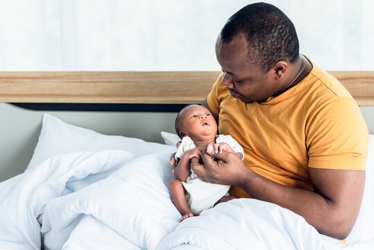 An African American father looking and kissing and holding hands  his 12-day-old baby newborn son lying in bed in a white bedroom, with happy, concept to African American family and newborn