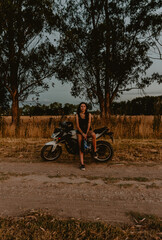 young woman looking at camera leaning on motorcycle, dirt road, background trees, golden hour
