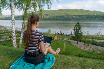 Girl is sitting on the bank of the river. In a lap netbook with a black screen. View from the back. Green grass in summer. Concept of remote study or work in nature.