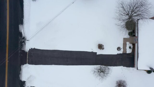 Top down aerial of snow covered home. Driveway to garage and house shoveled, cleared. Car drives on road. Winter, snow, holiday, travel theme.