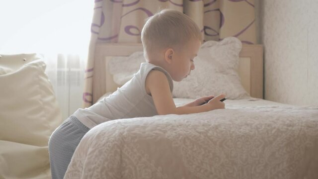 Little boy sitting on a bed and plays on the tablet in the room. Education and technology concept.