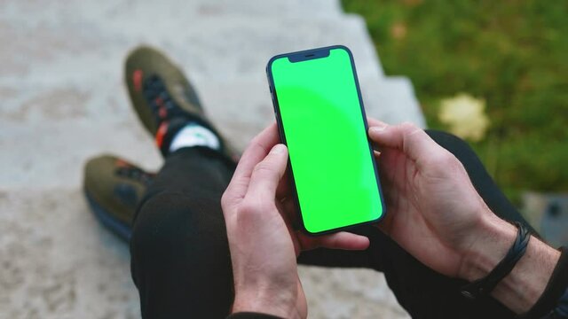Over the shoulder shot of man hands use phone with vertical green screen outdoors. Sitting. Touchscreen finger internet cellphone scrolling. Close up. Slow motion