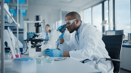 Medical Development Laboratory: Portrait of Black Male Scientist Looking Under Microscope, Analyzing Petri Dish Sample. Professionals Doing Research in Advanced Scientific Lab. Side View Shot - Powered by Adobe