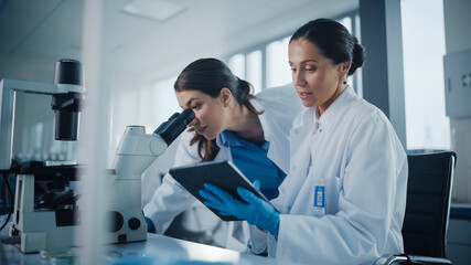 Modern Medical Research Laboratory: Two Female Scientists Working Together Using Microscope,...