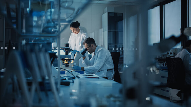 Modern Medical Research Laboratory: Two Scientists Working, Using Digital Tablet, Analyzing Test, Talking. Advanced Scientific Pharmaceutical Lab for Medicine, Biotechnology Development. Evening Time