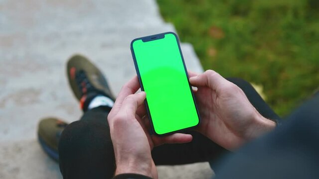 Close up over the shoulder shot of man hands use phone with vertical green screen outdoors. Sitting. Touchscreen finger internet cellphone scrolling. Slow motion