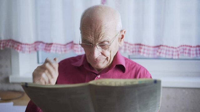 Old gray haired retired man holds eyeglasses with hand and reads newspaper close view