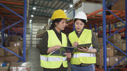 two asian female worker colleagues with clipboard and digital pad in warehouse discussing on new project. stockroom lady staffs in hardhat and safety vest working together. coworkers in storehouse.