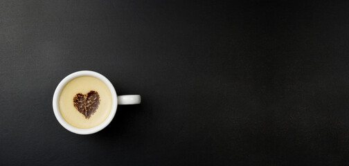 Cup of cappuccino in the center of the heart, on a black background with copy space. Chocolate heart in a cup with a hot coffee drink.