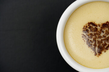 Close-up of cappuccino in the center of the heart, over black background with copy space. Chocolate heart in a mug with a hot coffee drink.