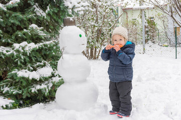 Happy child eating the carrot of the snowman while playing outdoor in winter. Cute little child building a snowman and having fun in park or backyard.