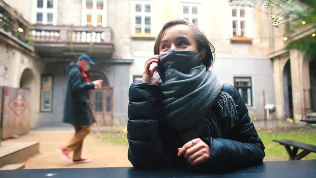 A woman in a mask sits at a table talking on the phone in a street cafe. Transmits a verbal message using the mobile Internet. A passer-by is moving in the background.