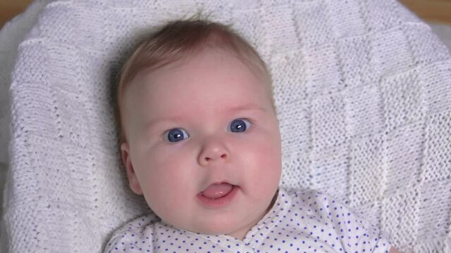 Happy cute blue-eyed baby is smiling and looking at the camera on the background of a white knitted blanket 
