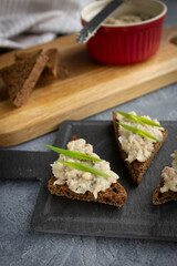 Canapes with herring and rye bread forshmak with green onions on a black tapas board. Background: ramekin with forshmak, slices of bread on a wooden board with a knife