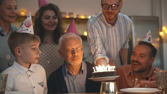 Gathering of friends and family at birthday party for senior man blowing out birthday candles.