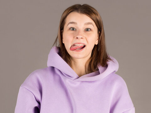 Funny cute young woman smiling showing tongue looking at camera over gray background.