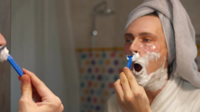 Young man with eye patches shaving face standing near mirror in modern bathroom at home in morning.