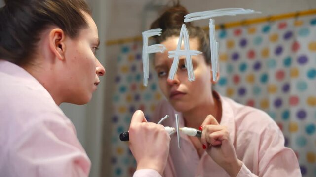 Young slim stressed woman writing fat word on bathroom mirror