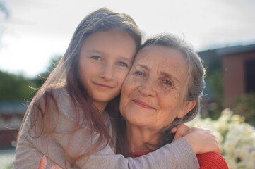 Senior grandmother with gray hair wearing red sweater with her little granddaughter are hugging in the garden and during sunny day outdoors, mother's day