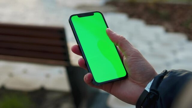 At sunlight young hands man holding use phone with vertical green screen in park background bench. Internet finger gadget device. Slow motion