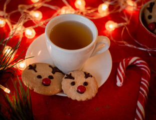 Christmas card, decor, cup of tea, gingerbread New Year's deer, candies, lights, tree on a red background