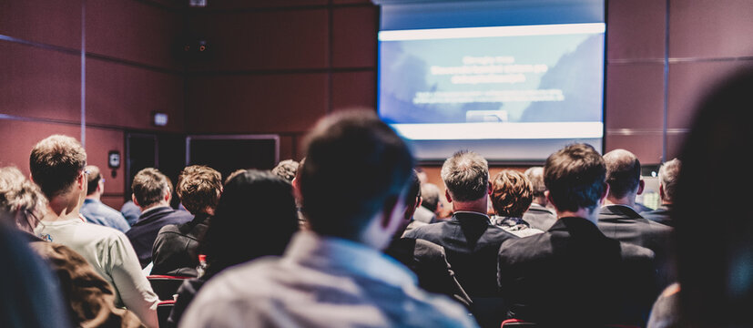 Business and entrepreneurship symposium. Speaker giving a talk at business meeting. Audience in conference hall. Rear view of unrecognized participant in audience.