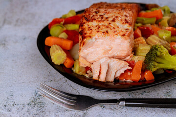 Baked juicy salmon fillet with steamed vegetables on black plate.
