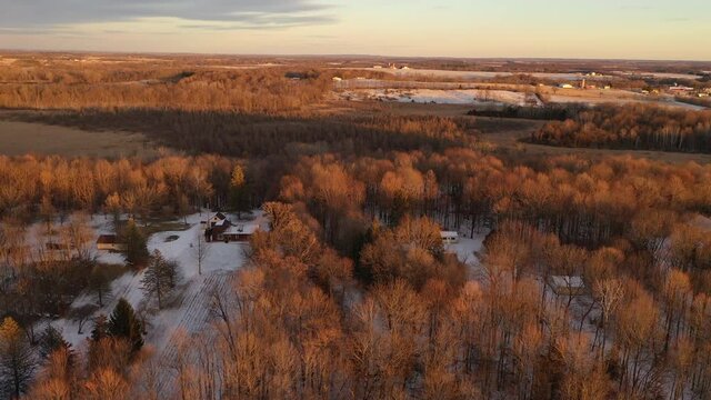 Establishing shot of Midwestern countryside in spring winter season. Aerial view of residential house, outbuildings in the forest. Sunny, soft sunlight at sunset