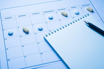 Taking pills due to time concept. Close up view photo of calendar with lying pills on the cells and open blank note pad with writing pen