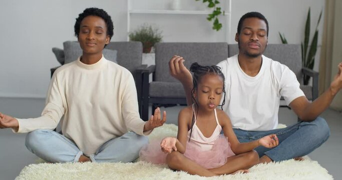 Calm afro american family mom dad and daughter threesome sit on floor in lotus position meditate together get rid of stress tension do yoga exercise develop spiritually mentally mindful life concept