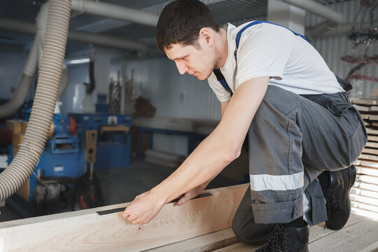 A young man works in a carpentry shop. An employee of Caucasian appearance in overalls measures wood with a ruler
