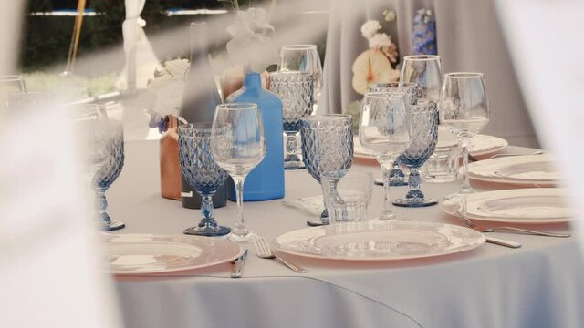 Table setting, beautiful clean and empty glass goblets, decorative bottles with flowers and pink colored large plates stand on a white tablecloth on a decorated table, man hand puts glass on table