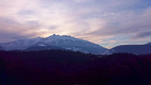 Drone aerial video footage above the Levico lake, facing the mountain Massiccio della vigolana, with sun setting in between the mountains behind the clouds. Flying above the water