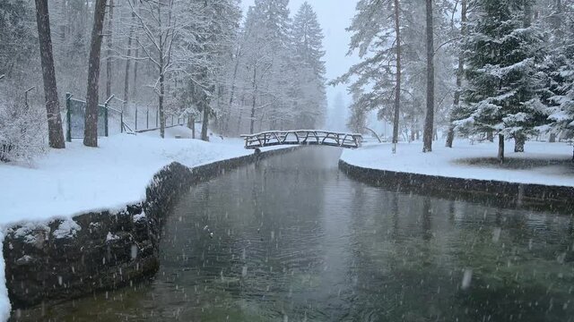 River flowing through stone canal with a wooden bridge in fairy tale winter landscape. Snowstorm in campground Sobec, Slovenia. Amazing view of snowflakes falling. Static shot, real time