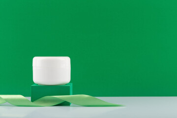 White jar with cream, mask or balm on green podium with ribbon on white glossy table against green background with copy space. Concept of organic cosmetics or daily skincare