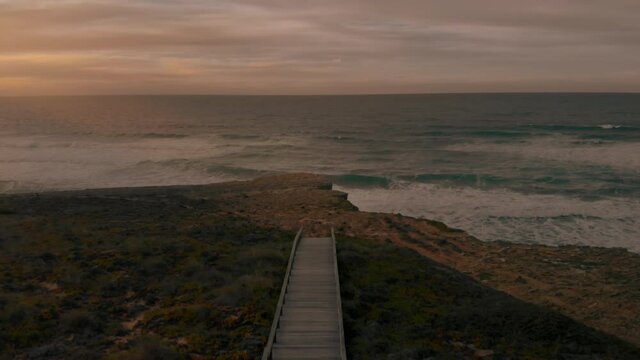 Cinematic drone shot of empty wooden boardwalk on rocky seaside. Road to nowhere, conceptual video of national park, path lead to ocean cliffs. Inspiring epic sunset shot of untouched nature