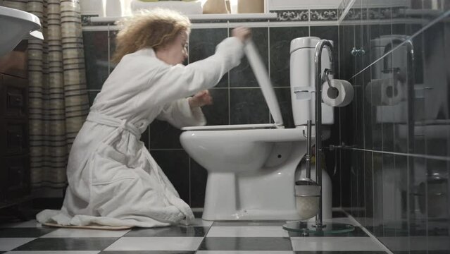 Side view of sick or pregnant woman opening wc and vomiting in toilet. Wide shot of unhealthy Caucasian lady in white bathrobe in bathroom. Intoxication or pregnancy concept.