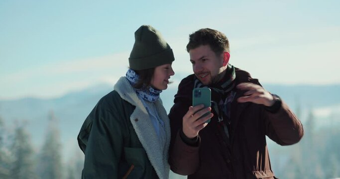 Affectionate young caucasian couple in warm outfits using smartphone taking pictures on mountain peak enjoying sunny weather during vacation.