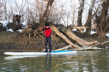 Senior paddler in a dry suit is training on stand up paddleboard on a Danube river in winter...