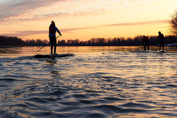 Silhouettes of three people on SUP (stand up paddle board) during a colorful sunset in the calm winter Danube river