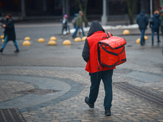 Obraz premium Delivery boy delivering online food orders to costumers, walking with red backpack, grocery delivery. Courier carries bag with fresh food. Delivering food at doorstep in holidays from restaurant