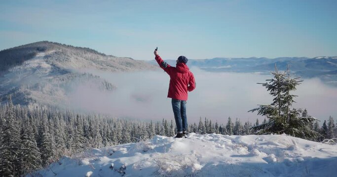 Joyful caucasian male traveler standing on top mountain taking selfie picture smartphone device enjoying cinematic view outdoors. Winter resort. Travel people.