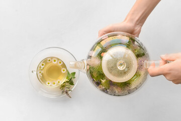 Woman pouring herbal tea from teapot in cup on light background
